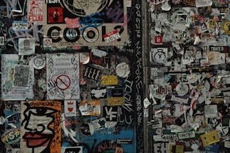 A dense collage of various stickers and graffiti covers a wall. The wall features a mix of colorful and monochrome stickers, tags, and small posters overlapping each other. Among the visual elements are bold text, stylized designs, and hand-drawn elements. Some stickers contain messages, logos, and artistic illustrations.