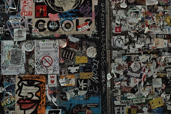 A dense collage of various stickers and graffiti covers a wall. The wall features a mix of colorful and monochrome stickers, tags, and small posters overlapping each other. Among the visual elements are bold text, stylized designs, and hand-drawn elements. Some stickers contain messages, logos, and artistic illustrations.