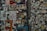 A dense collage of various stickers and graffiti covers a wall. The wall features a mix of colorful and monochrome stickers, tags, and small posters overlapping each other. Among the visual elements are bold text, stylized designs, and hand-drawn elements. Some stickers contain messages, logos, and artistic illustrations.