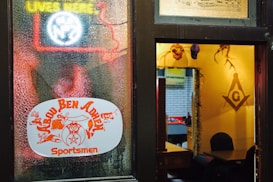 A colored glass door with a neon sign and stickers showing symbols and writing. The room inside has warm lighting with wall decorations including a prominent symbol resembling the Freemason emblem. There are helmets hung on the wall and a dim room beyond with more decorations.