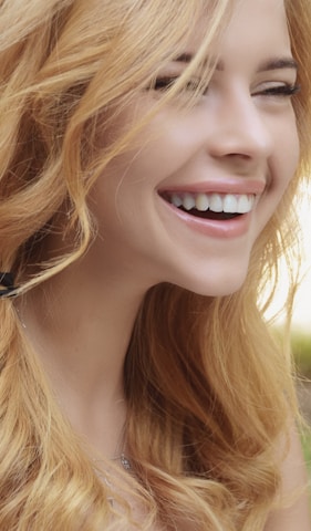 A woman with long blonde hair is smiling brightly. Her eyes are closed and she appears to be laughing or expressing joy. The focus is on her radiant smile and wavy hair.