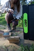A person kneeling on the ground in an outdoor setting, apparently working with electronic equipment. The individual is holding a device connected by red and black wires to a larger piece of equipment which appears to be a solar panel. There are open cardboard boxes on the ground around the person, likely containing components or accessories for the equipment.