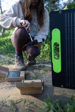 A person kneeling on the ground in an outdoor setting, apparently working with electronic equipment. The individual is holding a device connected by red and black wires to a larger piece of equipment which appears to be a solar panel. There are open cardboard boxes on the ground around the person, likely containing components or accessories for the equipment.
