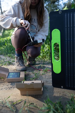 A person kneeling on the ground in an outdoor setting, apparently working with electronic equipment. The individual is holding a device connected by red and black wires to a larger piece of equipment which appears to be a solar panel. There are open cardboard boxes on the ground around the person, likely containing components or accessories for the equipment.
