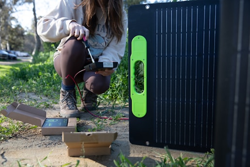 A person crouched on the ground, holding a device with wires connecting to a solar panel. The individual is wearing a sweater, leggings, and hiking boots, and the scene takes place outdoors in a grassy area. Small boxes are also present on the ground, likely containing equipment or tools.