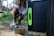 A person crouched on the ground, holding a device with wires connecting to a solar panel. The individual is wearing a sweater, leggings, and hiking boots, and the scene takes place outdoors in a grassy area. Small boxes are also present on the ground, likely containing equipment or tools.