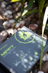 A solar charge controller with a green logo and text is placed on a surface covered with small pebbles. Sunlight casts shadows of nearby leaves onto the device, creating a pattern of light and shadow.