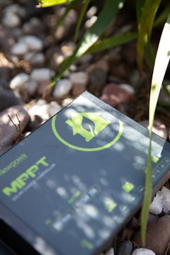 A solar charge controller with a green logo and text is placed on a surface covered with small pebbles. Sunlight casts shadows of nearby leaves onto the device, creating a pattern of light and shadow.