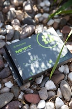 A Newpowa MPPT solar charge controller placed on a bed of variously colored pebbles with some green leaves partially covering it. The device is grey with green text and symbols, detailing specifications like PV 55V max and 12/24V 20A battery.