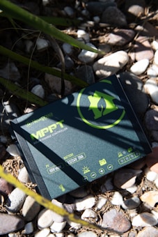 A solar charge controller device labeled 'MPPT' with a green eco-friendly design is resting on a bed of pebbles under the partial shade of green leaves. The device has specifications such as 55V max and 12/24V 20A printed on it.