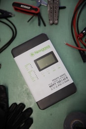 A solar charge controller from the Newpowa Frigate Series is situated on a green surface. Nearby are various tools including a multitool, pliers, a screwdriver set, and some cables. A pair of gloves and a roll of tape are also visible.
