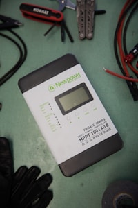 A solar charge controller from the Newpowa Frigate Series is situated on a green surface. Nearby are various tools including a multitool, pliers, a screwdriver set, and some cables. A pair of gloves and a roll of tape are also visible.