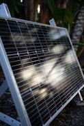 A solar panel is propped up outdoors, featuring reflective sunlight on its surface. The background consists of lush green foliage, indicating a garden or natural setting.