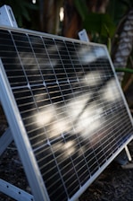 A solar panel is propped up outdoors, featuring reflective sunlight on its surface. The background consists of lush green foliage, indicating a garden or natural setting.