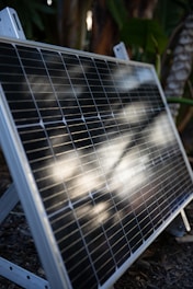 A solar panel is propped up outdoors, featuring reflective sunlight on its surface. The background consists of lush green foliage, indicating a garden or natural setting.