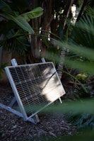 A solar panel is positioned on a wooden or mulch-covered ground within a lush garden setting. Surrounding the panel are various tropical plants with large, green leaves. The sunlight reflects on the solar panel, highlighting its grid pattern against the backdrop of dense foliage.