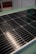 A close-up view of a solar panel with rows of rectangular photovoltaic cells. The surface appears glossy and reflective, indicating a clean and new condition. The background is slightly blurred, focusing attention on the panel's details.