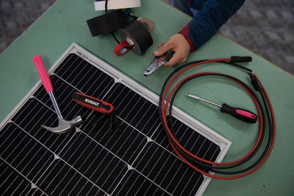 A collection of tools and materials on a flat surface, including a solar panel, a hammer with a pink handle, a multi-tool labeled 'Kobalt', an insulated screwdriver, and a pair of cables with connectors. A hand holds a wire cutter near several rolls of electrical tape.