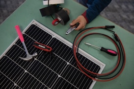 A collection of tools and materials on a flat surface, including a solar panel, a hammer with a pink handle, a multi-tool labeled 'Kobalt', an insulated screwdriver, and a pair of cables with connectors. A hand holds a wire cutter near several rolls of electrical tape.