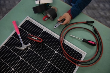 A collection of tools and materials on a flat surface, including a solar panel, a hammer with a pink handle, a multi-tool labeled 'Kobalt', an insulated screwdriver, and a pair of cables with connectors. A hand holds a wire cutter near several rolls of electrical tape.