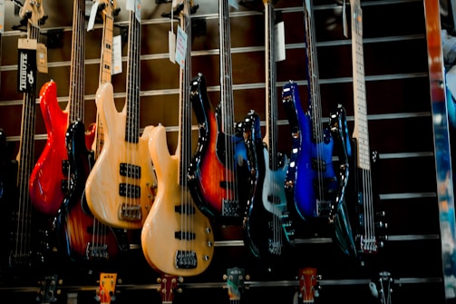 A display of various electric bass guitars in a music store. The guitars are arranged in a row, showcasing different colors and designs, including red, natural wood, sunburst, and blue. They are hung on a slatted wall, and each guitar has a tag hanging from its tuning pegs.