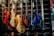 A display of various electric bass guitars in a music store. The guitars are arranged in a row, showcasing different colors and designs, including red, natural wood, sunburst, and blue. They are hung on a slatted wall, and each guitar has a tag hanging from its tuning pegs.