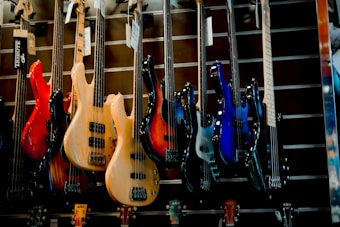 A display of various electric bass guitars in a music store. The guitars are arranged in a row, showcasing different colors and designs, including red, natural wood, sunburst, and blue. They are hung on a slatted wall, and each guitar has a tag hanging from its tuning pegs.