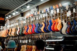 A music store showcasing a wide array of electric guitars and a keyboard. The guitars, displayed on the wall, are in a variety of vivid colors including blue, red, orange, and natural wood finishes. The store is well-lit, highlighting the glossy finishes of the instruments. Ceiling lights and branding signs are visible, adding to the commercial music shop setting.