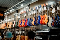 A music store showcasing a wide array of electric guitars and a keyboard. The guitars, displayed on the wall, are in a variety of vivid colors including blue, red, orange, and natural wood finishes. The store is well-lit, highlighting the glossy finishes of the instruments. Ceiling lights and branding signs are visible, adding to the commercial music shop setting.