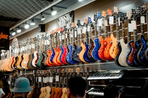 A music store showcasing a wide array of electric guitars and a keyboard. The guitars, displayed on the wall, are in a variety of vivid colors including blue, red, orange, and natural wood finishes. The store is well-lit, highlighting the glossy finishes of the instruments. Ceiling lights and branding signs are visible, adding to the commercial music shop setting.