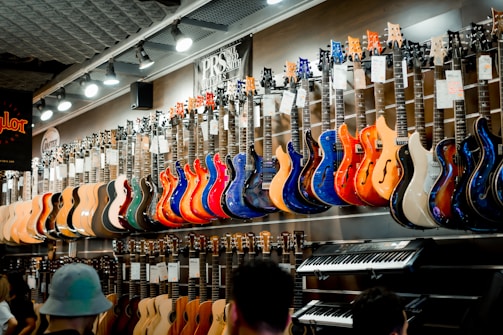 A music store showcasing a wide array of electric guitars and a keyboard. The guitars, displayed on the wall, are in a variety of vivid colors including blue, red, orange, and natural wood finishes. The store is well-lit, highlighting the glossy finishes of the instruments. Ceiling lights and branding signs are visible, adding to the commercial music shop setting.