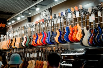A music store showcasing a wide array of electric guitars and a keyboard. The guitars, displayed on the wall, are in a variety of vivid colors including blue, red, orange, and natural wood finishes. The store is well-lit, highlighting the glossy finishes of the instruments. Ceiling lights and branding signs are visible, adding to the commercial music shop setting.