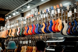 A music store showcasing a wide array of electric guitars and a keyboard. The guitars, displayed on the wall, are in a variety of vivid colors including blue, red, orange, and natural wood finishes. The store is well-lit, highlighting the glossy finishes of the instruments. Ceiling lights and branding signs are visible, adding to the commercial music shop setting.