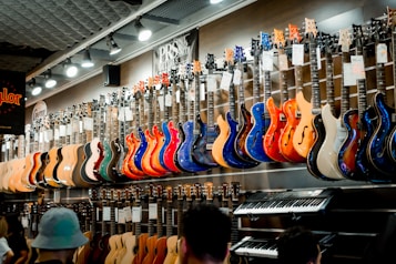 A music store showcasing a wide array of electric guitars and a keyboard. The guitars, displayed on the wall, are in a variety of vivid colors including blue, red, orange, and natural wood finishes. The store is well-lit, highlighting the glossy finishes of the instruments. Ceiling lights and branding signs are visible, adding to the commercial music shop setting.