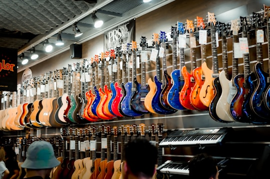 A music store showcasing a wide array of electric guitars and a keyboard. The guitars, displayed on the wall, are in a variety of vivid colors including blue, red, orange, and natural wood finishes. The store is well-lit, highlighting the glossy finishes of the instruments. Ceiling lights and branding signs are visible, adding to the commercial music shop setting.