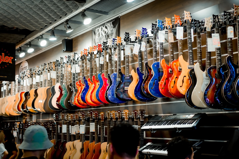 A music store showcasing a wide array of electric guitars and a keyboard. The guitars, displayed on the wall, are in a variety of vivid colors including blue, red, orange, and natural wood finishes. The store is well-lit, highlighting the glossy finishes of the instruments. Ceiling lights and branding signs are visible, adding to the commercial music shop setting.