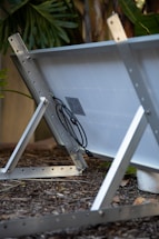 A solar panel is mounted on a metal frame, angled on a bed of mulch with greenery in the background. The panel's connectors and wiring are visible, attached to the back.