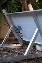 A solar panel is mounted on a metal frame, angled on a bed of mulch with greenery in the background. The panel's connectors and wiring are visible, attached to the back.