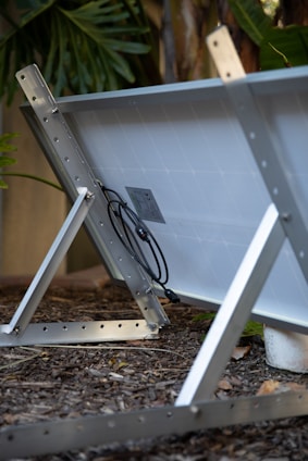 A solar panel is mounted on a metal frame, angled on a bed of mulch with greenery in the background. The panel's connectors and wiring are visible, attached to the back.