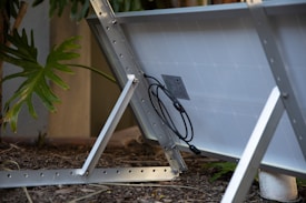 A solar panel is positioned at an angle with visible support structures and electrical cables connected at the back. The panel is situated on mulch-covered ground, near a building wall with large green leaves partially visible, indicating a garden or natural setting.