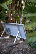 A solar panel placed on the ground supported by metal frames in a lush garden with various green plants and trees surrounding it.
