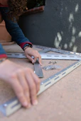A person with curly hair is assembling metal brackets on a concrete surface. They are using their hands to align the pieces, and small hardware like screws and bolts are visible in a plastic bag nearby. The person is wearing a blue long-sleeve shirt and a smartwatch.