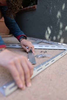 A person with curly hair is assembling metal brackets on a concrete surface. They are using their hands to align the pieces, and small hardware like screws and bolts are visible in a plastic bag nearby. The person is wearing a blue long-sleeve shirt and a smartwatch.