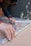 A person with curly hair is assembling metal brackets on a concrete surface. They are using their hands to align the pieces, and small hardware like screws and bolts are visible in a plastic bag nearby. The person is wearing a blue long-sleeve shirt and a smartwatch.
