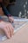 A person with curly hair is assembling metal brackets on a concrete surface. They are using their hands to align the pieces, and small hardware like screws and bolts are visible in a plastic bag nearby. The person is wearing a blue long-sleeve shirt and a smartwatch.