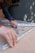 A person with curly hair is assembling metal brackets on a concrete surface. They are using their hands to align the pieces, and small hardware like screws and bolts are visible in a plastic bag nearby. The person is wearing a blue long-sleeve shirt and a smartwatch.