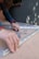 A person with curly hair is assembling metal brackets on a concrete surface. They are using their hands to align the pieces, and small hardware like screws and bolts are visible in a plastic bag nearby. The person is wearing a blue long-sleeve shirt and a smartwatch.