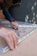 A person with curly hair is assembling metal brackets on a concrete surface. They are using their hands to align the pieces, and small hardware like screws and bolts are visible in a plastic bag nearby. The person is wearing a blue long-sleeve shirt and a smartwatch.