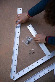 Two hands are assembling metal beams with holes on a rough surface. There is a small plastic bag containing bolts and washers next to the beams.