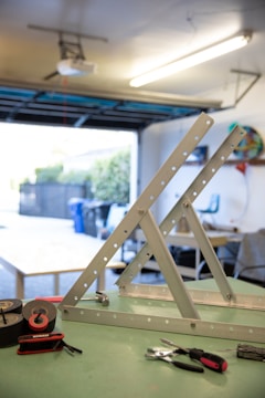 A workspace in what appears to be a garage, where metal frameworks and various tools are arranged on a green tabletop. Visible tools include electrical tape, pliers, and a screwdriver. The background shows a partially open garage door with some outdoor elements.